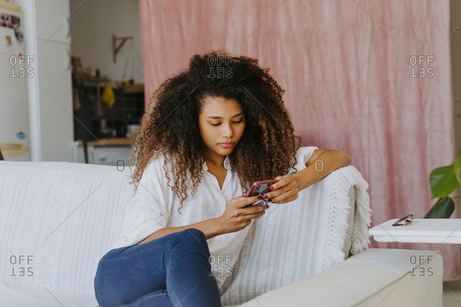 Young African American female in casual outfit sitting on sofa and surfing internet on mobile phone while resting in living room at home