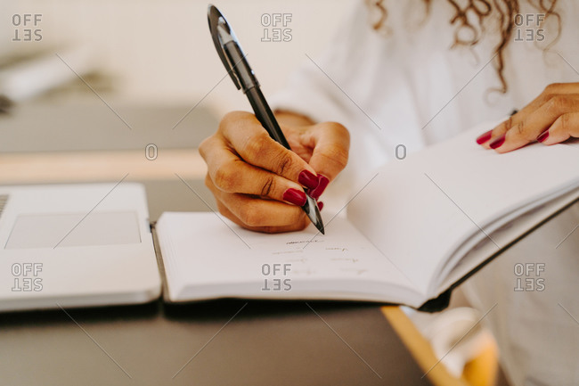 Crop unrecognizable female entrepreneur sitting at table with laptop and writing plan for business project in notebook