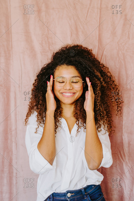 Attractive African American female with curly hair and in casual outfit standing on pink background and smiling while touching glasses
