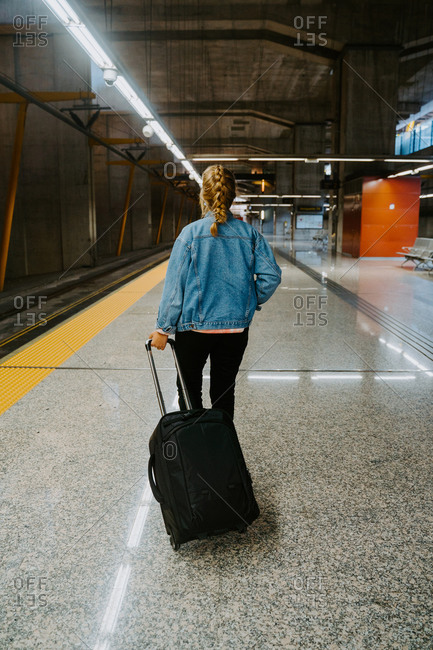 Full body back view of unrecognizable young female traveler wearing casual clothes walking with suitcase on railway station looking away