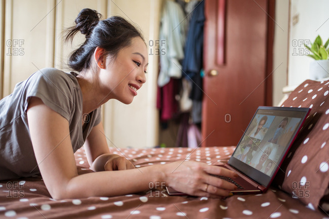 Side view of young Asian female lying on bed and chatting via video call on laptop with group of friends while spending time at home during coronavirus pandemic