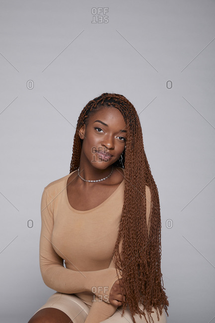 Relaxed African American female in casual outfit and with braids sitting on gray background in studio and tenderly looking at camera