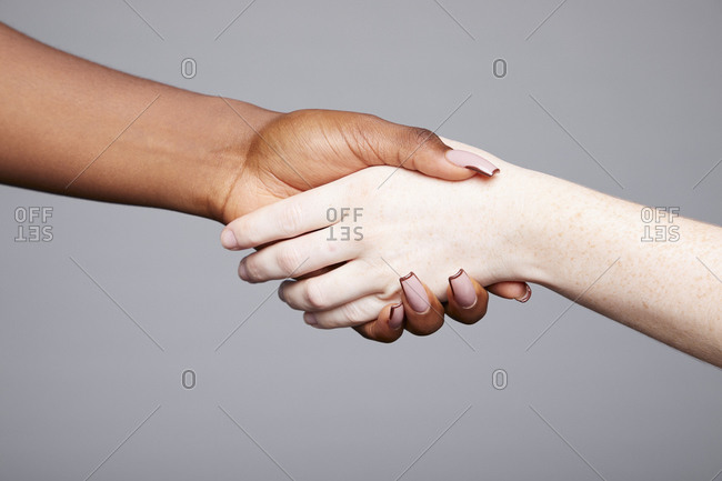 Crop unrecognizable multiethnic females holding and shaking hands as greeting and equal gesture on gray background in studio