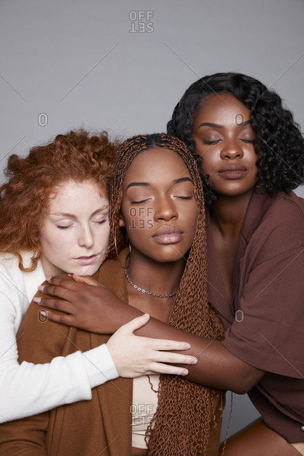 Tranquil multiracial females with braids and curly hair cuddling tenderly on gray background in studio with eyes closed