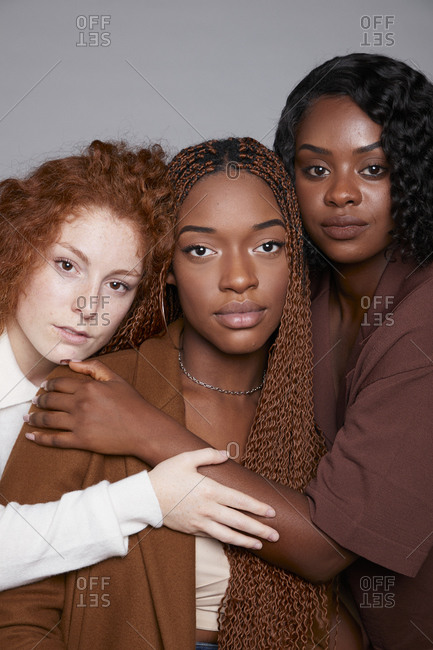 Tranquil multiracial females with braids and curly hair cuddling tenderly on gray background in studio looking at camera