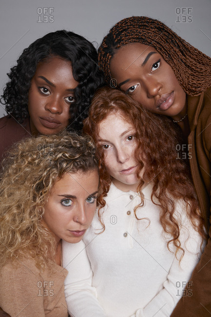 Tranquil multiracial females with braids and curly hair cuddling tenderly on gray background in studio looking at camera
