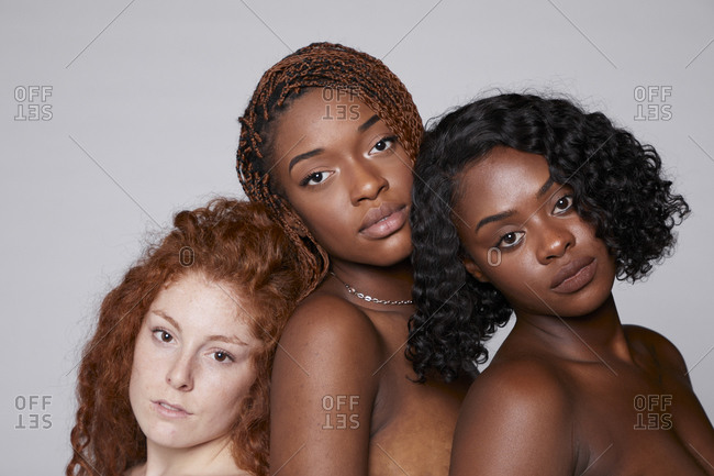 Tranquil multiracial females with bare shoulders cuddling tenderly on gray background in studio looking at camera