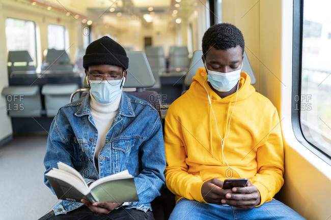 Busy African American male travelers in medical masks reading book and listening to music while sitting on passenger seats in train and entertaining during trip