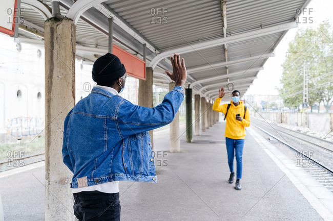 Black male friends in medical masks waving hands and greeting each other on train platform during coronavirus epidemic