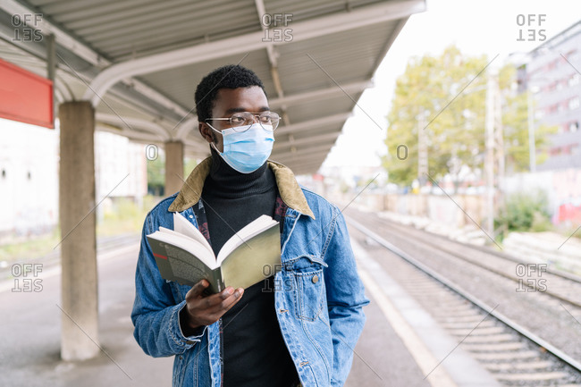 African American male traveler in medical mask during coronavirus pandemic standing on railroad platform with book while waiting for train and looking away