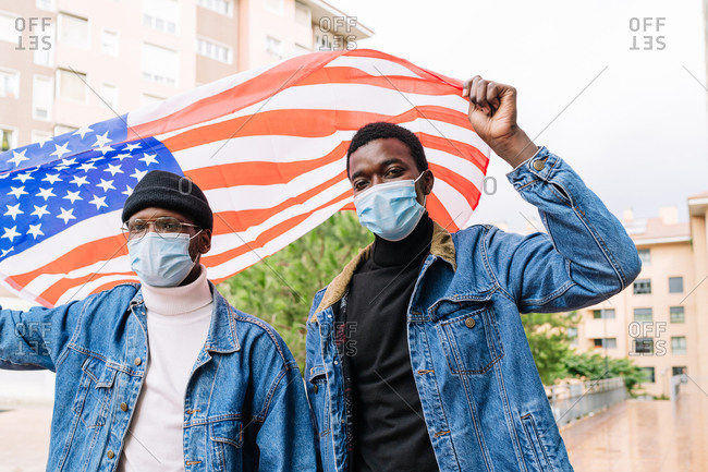 Low angle of cheerful African American male friends in masks standing with USA national flag on street and expressing joy