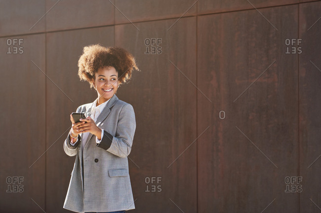 Positive ethnic female entrepreneur in smart casual style using smartphone while standing on street looking away