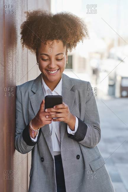 Positive ethnic female entrepreneur in smart casual style using smartphone while standing on street