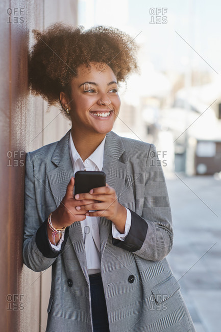 Positive ethnic female entrepreneur in smart casual style using smartphone while standing on street looking away
