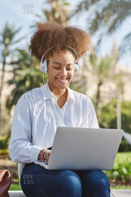 Positive African American businesswoman sitting on bench near suitcase and typing on laptop while working on remote project