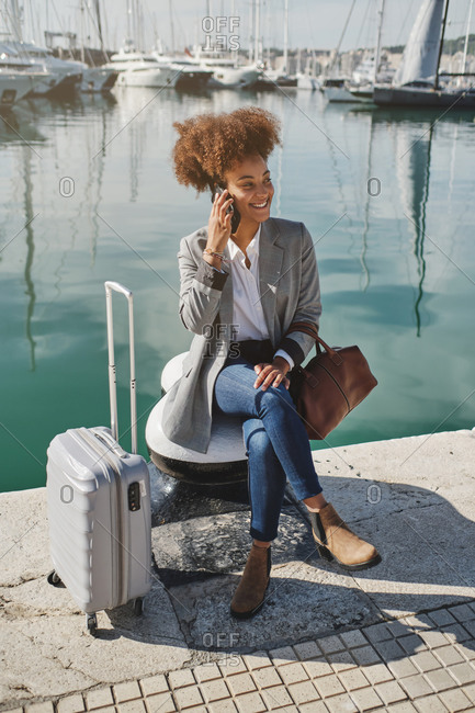 High angle of black businesswoman sitting on embankment with suitcase and discussing project on mobile phone