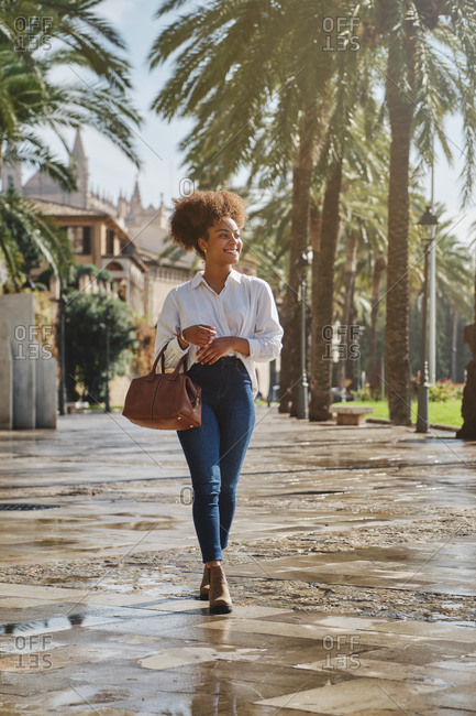 Cheerful African American female in casual outfit and with handbag walking along street in tropical city and looking away