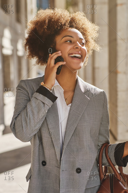 Delighted African American female in smart casual style walking in city on sunny day and speaking with friend on cellphone