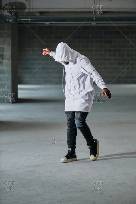 Full body of African American male in street style clothes and hood standing with outstretched arms in bright underground and looking down