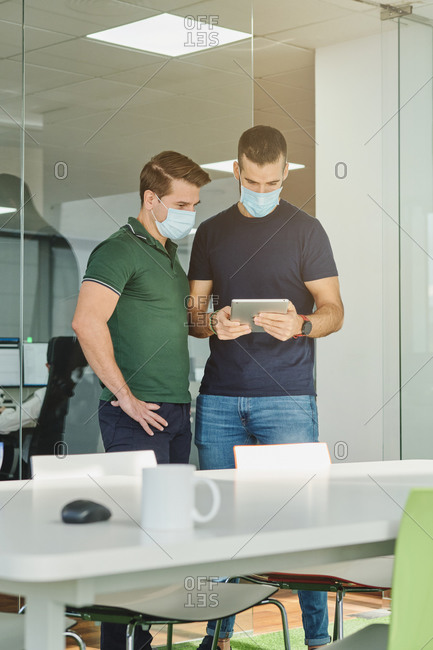 Busy male programmers in medical masks standing with tablet in office and discussing software development at work during coronavirus outbreak