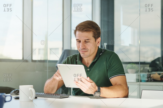 Concentrated male software developer sitting at table in modern workplace and browsing tablet while working on program code