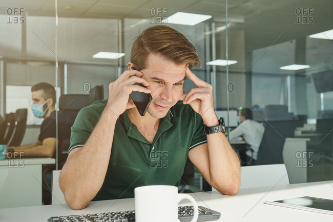 Smiling male employee sitting at table in workplace and discussing project on cellphone at working day
