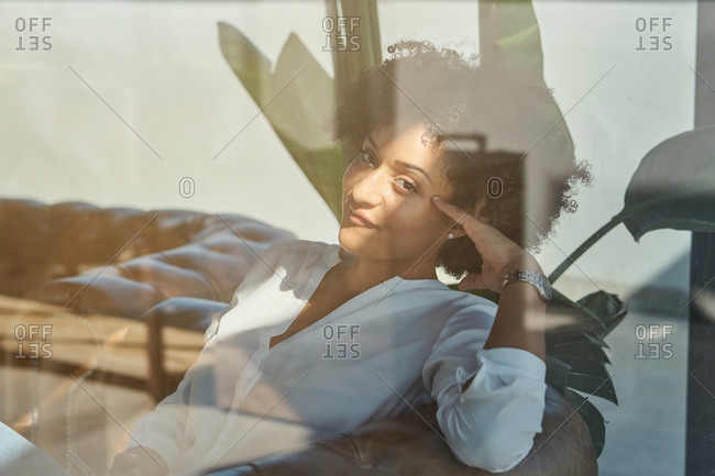 Tranquil African American female with curly hair leaning on hand and resting in comfortable armchair while tenderly looking at camera through glass