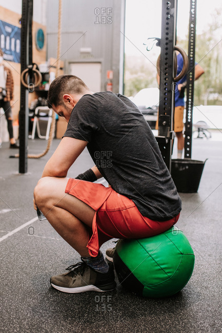 Side view of muscular male athlete sitting on medicine ball and having break during active training in sports center