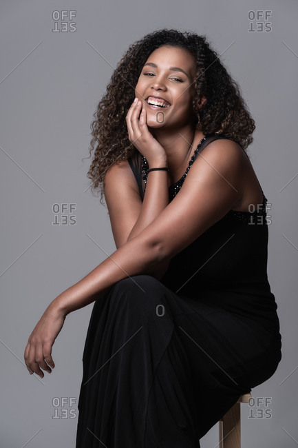 Cheerful young plus size African American female model with long curly hair wearing elegant black dress sitting on chair and looking at camera against gray background