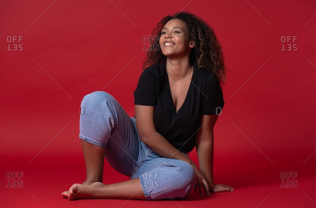 Full body of happy young overweight African American woman in casual black shirt and jeans with bare feet sitting in red studio