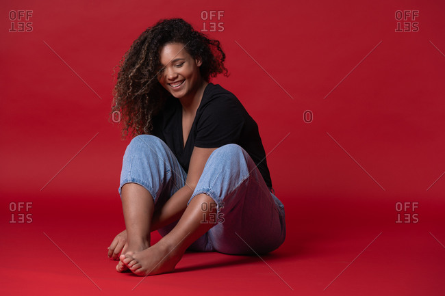 Full body of happy young overweight African American woman in casual black shirt and jeans with bare feet sitting in red studio