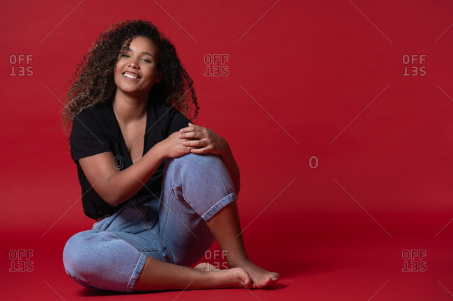 Full body of happy young overweight African American woman in casual black shirt and jeans with bare feet sitting in red studio looking at camera