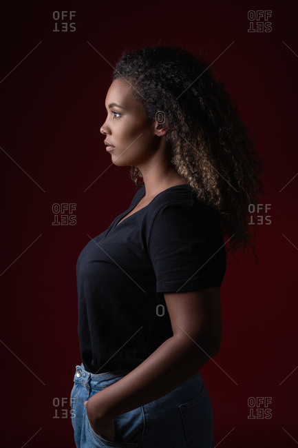 Side view of young overweight African American woman in casual black shirt and jeans standing in red studio looking away
