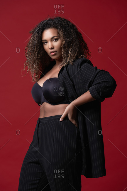 Beautiful confident young plus size black female model with long curly hair wearing elegant black dress looking at camera against red background
