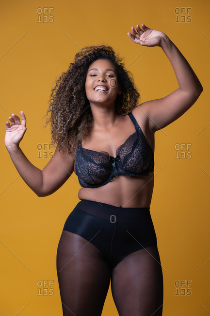 Plus size cheerful African American female model with long curly hair wearing elegant lace underwear looking at camera against yellow background