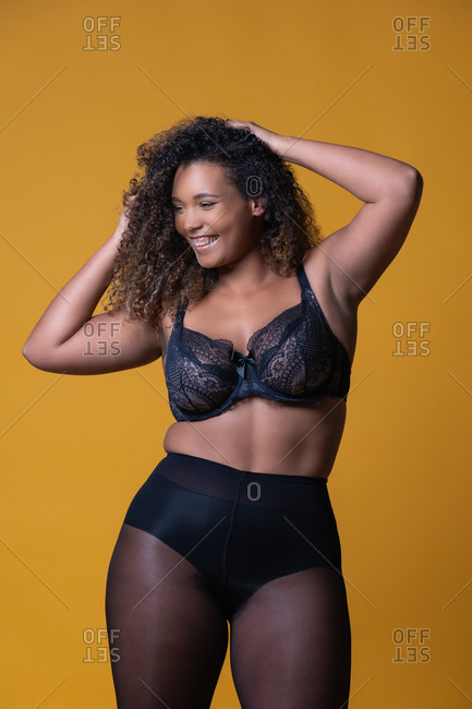 Plus size cheerful African American female model with long curly hair wearing elegant lace underwear looking at camera against yellow background