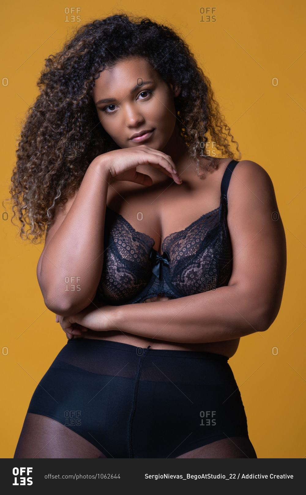 Attractive African American overweight female model with long curly hair  wearing delicate lace bra looking at camera against yellow background stock  photo - OFFSET