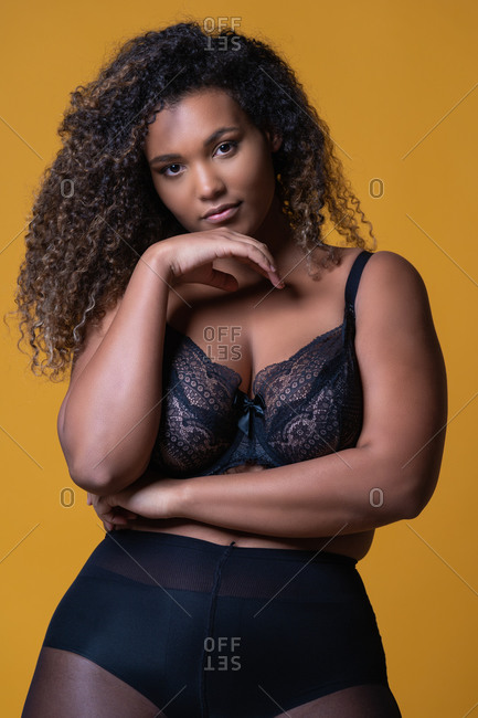 Attractive African American overweight female model with long curly hair wearing delicate lace bra looking at camera against yellow background