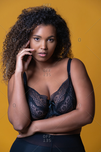 Attractive African American overweight female model with long curly hair wearing delicate lace bra looking at camera against yellow background