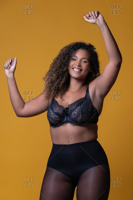 Plus size cheerful African American female model with long curly hair  wearing elegant lace underwear looking at camera against yellow background  stock photo - OFFSET