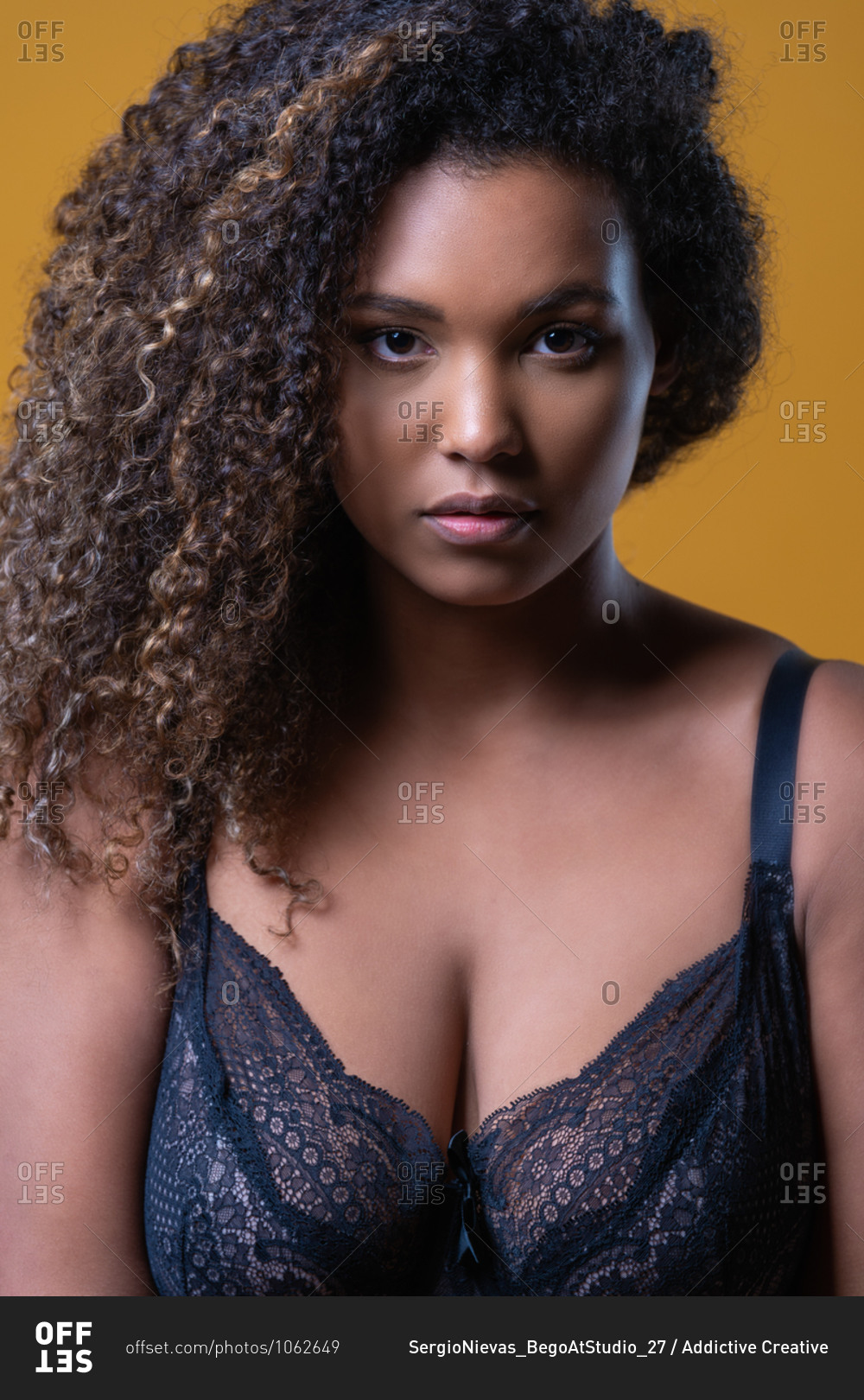 Attractive African American overweight female model with long curly hair  wearing delicate lace bra looking at camera against yellow background stock  photo - OFFSET