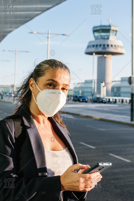 Traveling female wearing protective respirator during coronavirus epidemic browsing smartphone in airport after arrival and waiting for taxi