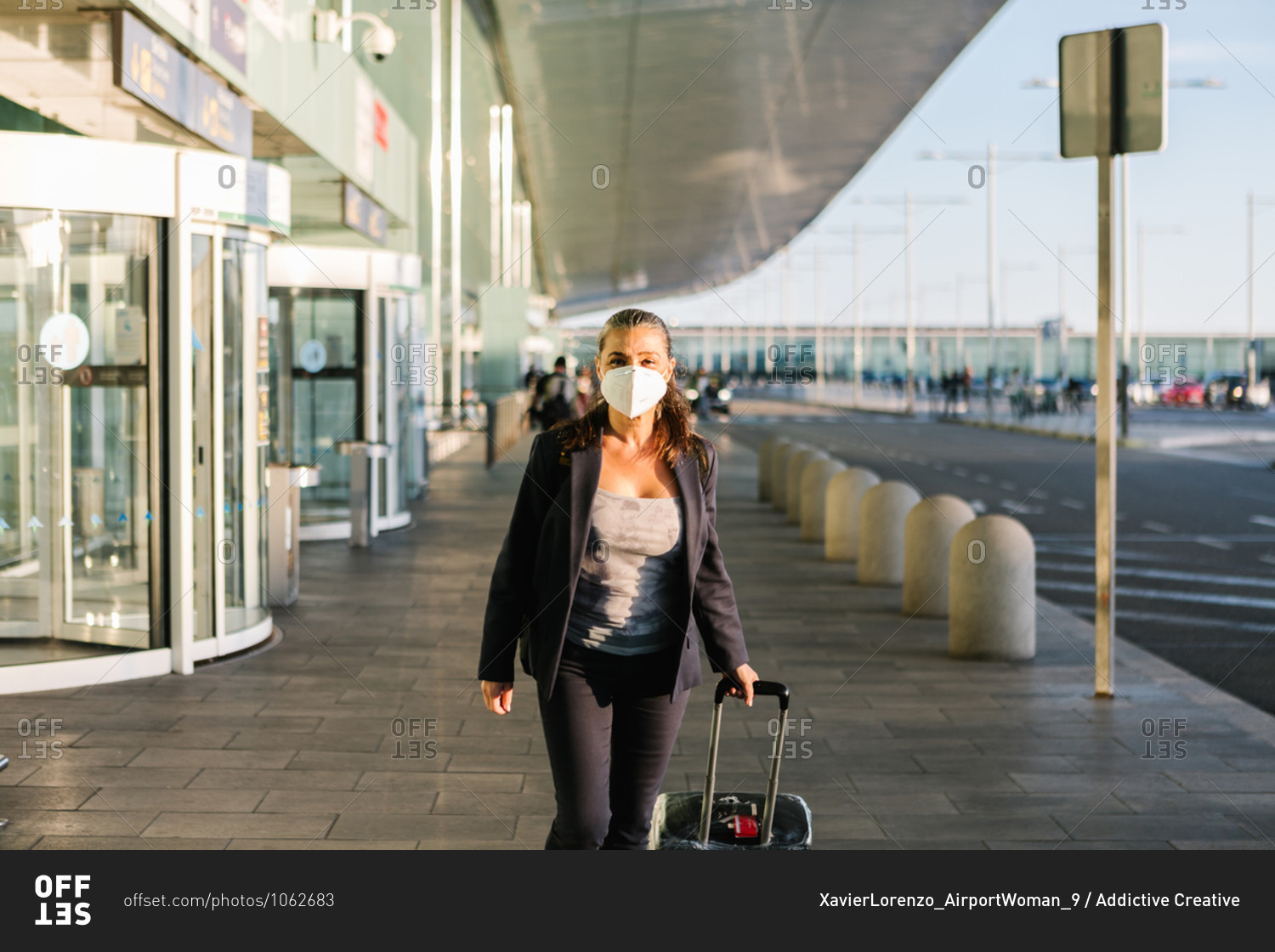 Cheerful female tourist in mask with suitcase and backpack standing in airport and smiling at camera while enjoying summer weekend