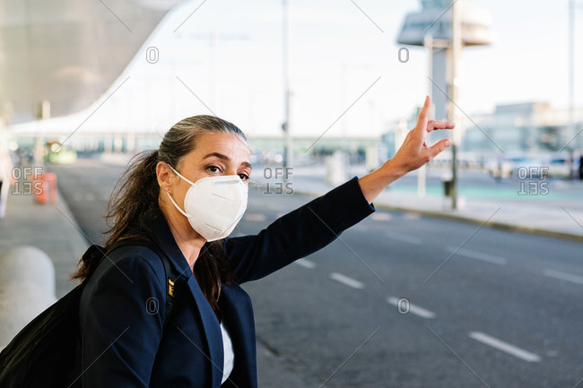 Female tourist in protective respirator during COVID 19 epidemic standing in airport and catching taxi with outstretched arm while looking away