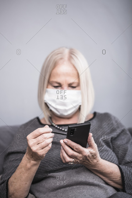 An elderly woman texting on her cell phone with a stylus pen while wearing a facemask