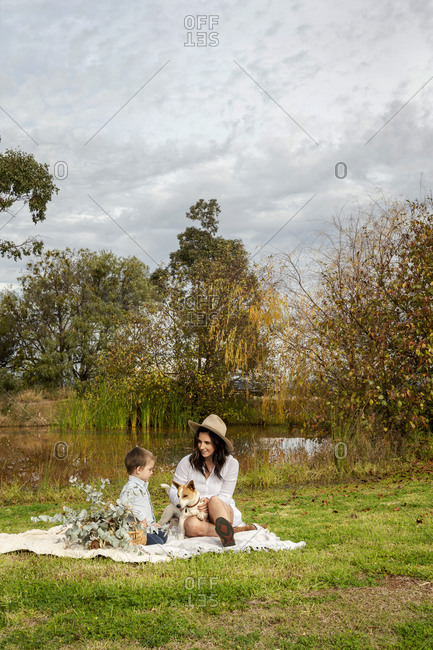 Mother and toddler boy sitting with dog on a picnic blanket beside pond