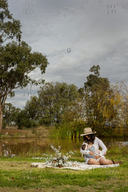 Mother breastfeeding her baby on a picnic blanket beside pond