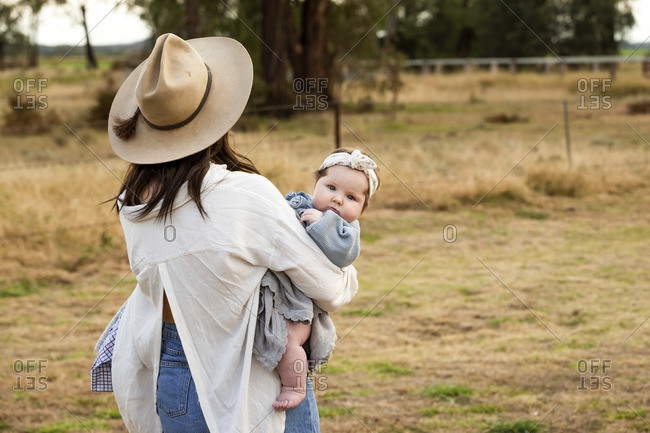 Mother wearing a cowgirl hat carrying her baby girl in a field
