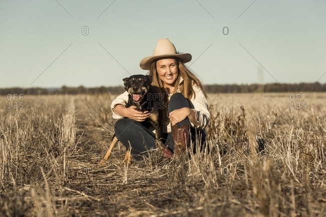 Portrait of a young blonde woman wearing a cowgirl hat sitting in a field with her dog
