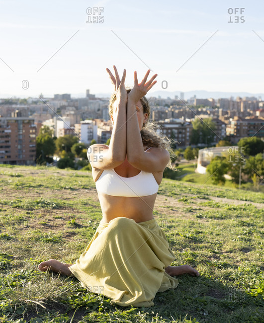 Full body unrecognizable barefoot female sitting in Cow Face Pose and hiding face behind arms while practicing yoga on sunny hill against city landscape